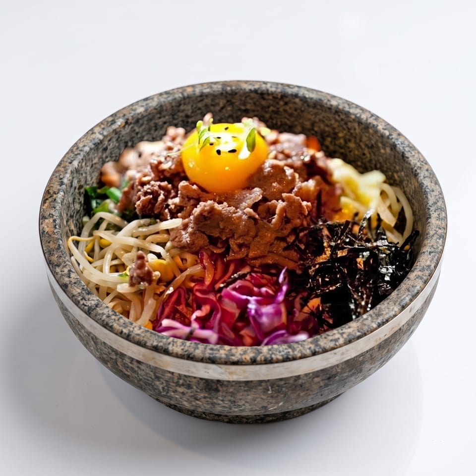 Colorful Korean rice bowl topped with crispy meat, vegetables, seaweed, and a bright egg yolk in a stone bowl.